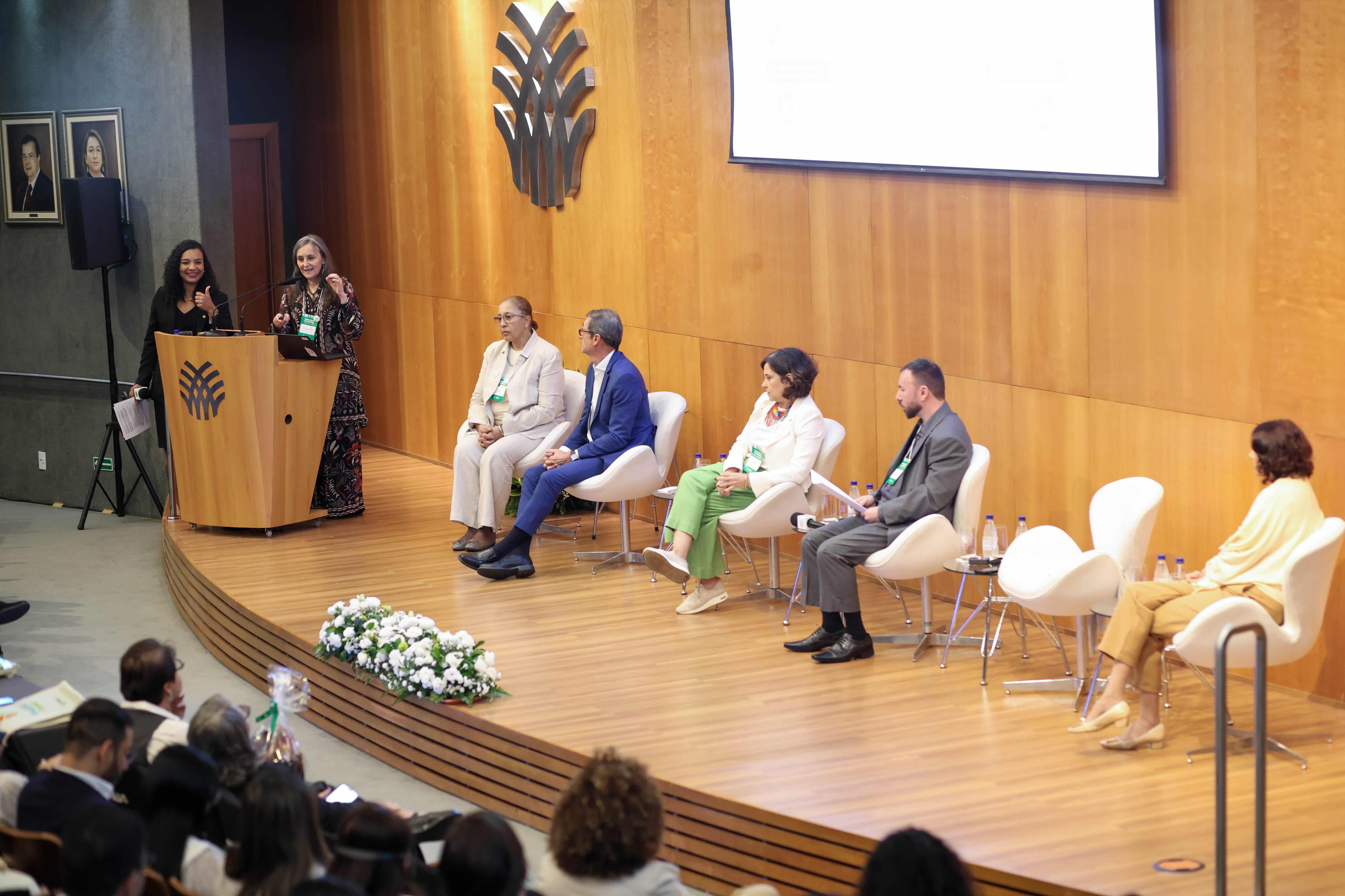 A Prof.ª Viviana Martinez-Bianchi fala no púlpito ao lado de uma colega, enquanto o painel do encontro está sentado no palco.